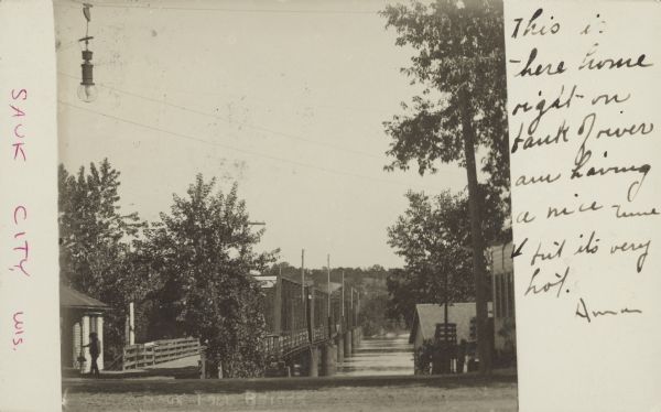 Written faintly along bottom: "Sauk Toll Bridge". The view is along the side of the bridge, with buildings and trees on both sides in the foreground. A man is walking in front of the building on the left. The support structure for the bridge is at the waterline. Trees and hills are on the far shoreline.