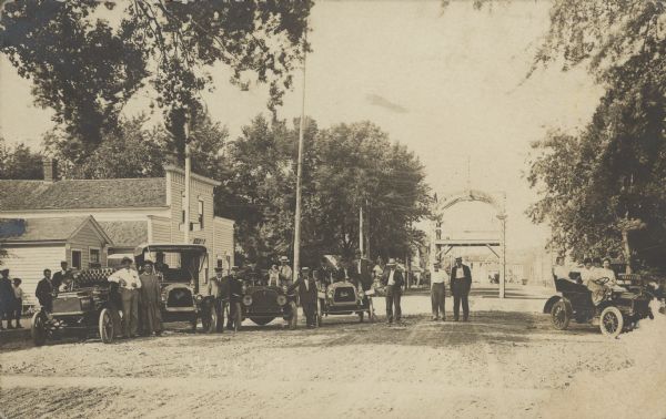 Faint handwriting across foot reads: "Sauk City, Wis." A group of people and their automobiles are posing in the unpaved road. An arched structure of some kind is in the center of the road behind them. Several buildings are on the left, and trees are on both sides of the road.