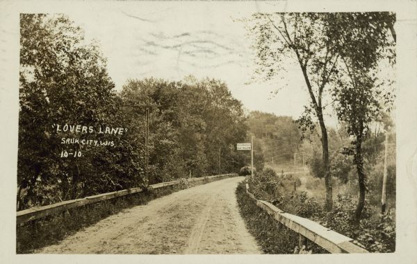 Text on front reads: "Lovers Lane, Sauk City, Wis." An unpaved, rural road with a wooden guard rail on both sides. A sign reads: "Automobiles Go Slow." Behind the sign is a farmer driving a horse-drawn hay wagon with a full load of hay. Many trees are on both sides and on the hills in the background.