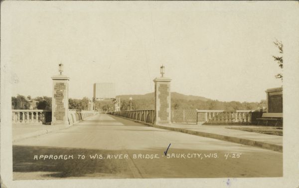 Text on front reads: "Approach to Wisconsin River Bridge - Sauk City, Wis." The view is straight across to the opposite side. Trees and hills are on the far side of the river. Two stone columns with lanterns on the top frame the entrance to the bridge, and stone balustrades are on both sides. Lampposts line both sides of the bridge, and a walkway is along the right side. On the far right is a bench in front of a brick wall with a plaque.