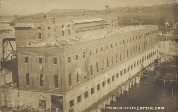 Text on front reads: "Power House at the Dam." Elevated view of the powerhouse, which is a very large brick and concrete structure next to the Prairie du Sac Hydroelectric Dam on the Wisconsin River. The dam began generating power in 1914. Lake Wisconsin is the result of damming the Wisconsin River. Men are working on the roof of the building.