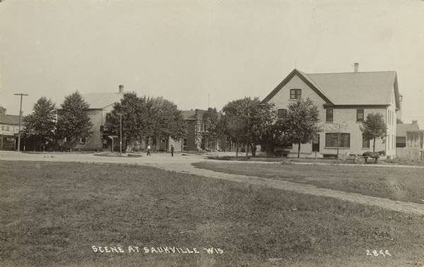Text on front reads: "Scene at Saukville, Wis." Small town scene of a neighborhood from a grassy field. Several dwellings are in the middle ground with commercial buildings behind them. Two men are standing in the street and a horse-drawn wagon is next to the house on the right. Several unpaved streets come together in a roundabout.