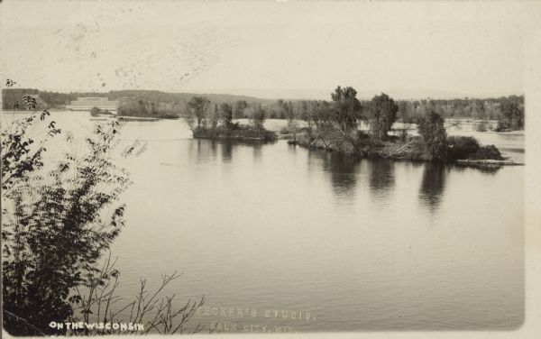 Text on front reads: "On the Wisconsin." Wooded islands on the Wisconsin River as seen from the shore. The opposite shoreline is in the background. The powerhouse for the Prairie du Sac Hydroelectric Dam can be seen in the distance on the left. This body of water is the beginning of Lake Wisconsin.