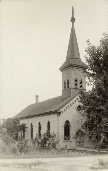 Text on front reads: "Saukville, Wis." The Evangelical Saint Peter's Church was built of cream brick with a clapboard belfry in 1876. The plaque above the front door reads: "EV ST PETERS CHURCH 1876." The church is surrounded by trees. The road in front is unpaved.