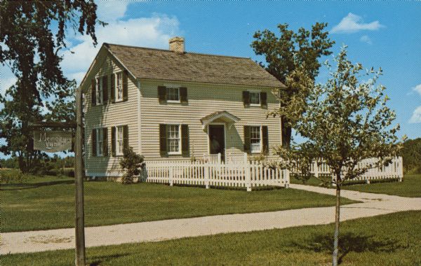A clapboard house with a white picket fence in the Ozaukee County Pioneer Village. Text on back reads: "Ozaukee County, Pioneer Village, Wisconsin. Halpin Cottage 1850's. Pre-Civil War frame cottage built in Five Corners north of Cedarburg by Patrick Halpin, an Irish immigrant. Designed with two rooms on each of the two floors. An addition made in 1864, containing a kitchen and bedroom, was removed during restoration. Square nails were used throughout. Exterior sheathing is fastened directly to the frame." A sign on the left reads: "Memory Lane" and mature trees are in the background.