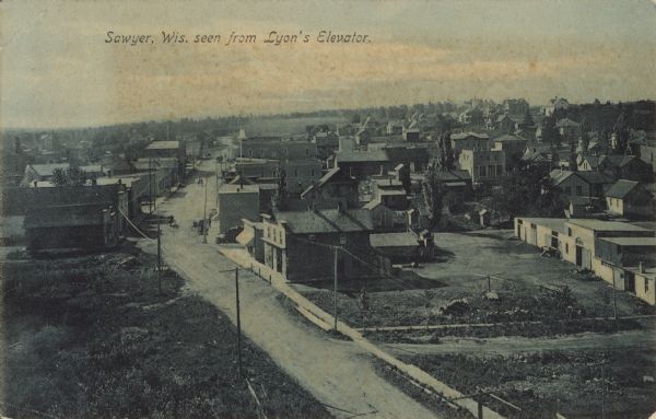 Text on front reads: "Sawyer, Wis. seen from Lyon's Elevator." An elevated view of the town from a grain elevator, with many homes and businesses. The street is unpaved and there are sidewalks on each side.