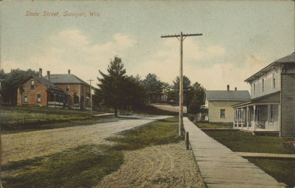 Text on front reads: "State Street, Sawyer, Wis." A residential unpaved street with dwellings on both sides.There is a wooden boardwalk on the right.