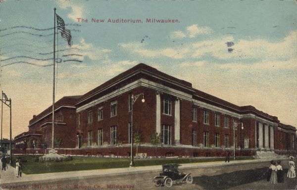 Text on front reads: "The New Auditorium, Milwaukee." A large brick building with stone trim, surrounded by a large lawn and sidewalks. Several people are strolling about and an automobile is on the street. An American flag flies from a pole on the corner.
