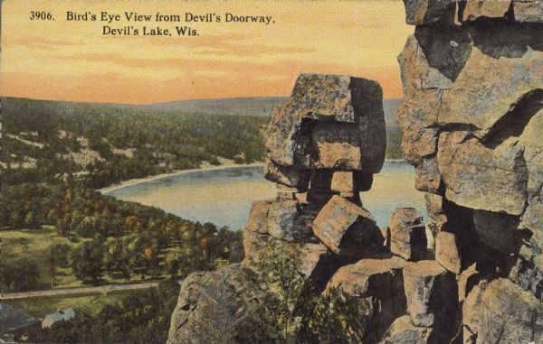 Text on front reads: "Bird's Eye View from Devil's Doorway. Devil's Lake, Wis." Elevated view of the rock formation called the Devil's Doorway in Devil's Lake State Park. Trees can be seen below. The lake, bluffs, roads and buildings are on to the left. The sky is orange.