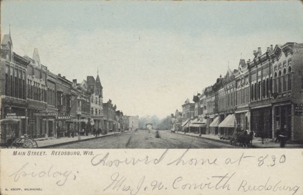 Text on front reads: "Main Street, Reedsburg, Wis." Street level view of the central business district. People are walking on the sidewalks and horse-drawn vehicles are in the unpaved street or hitched at the curb. Many business have signs and awnings, "Printing Office," "Millinery" and "Market."