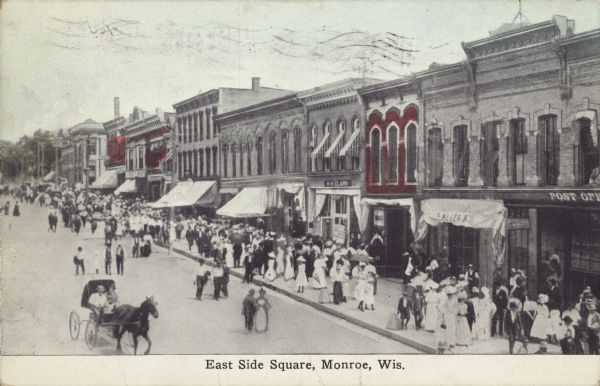 Caption reads: "East Side Square, Monroe, Wis." Elevated view of people walking on the sidewalks and in the street, and a horse and buggy driving by on the left. Many businesses have awnings, and some of the signs on the buildings read: "Post Office" and "S Miller 8."