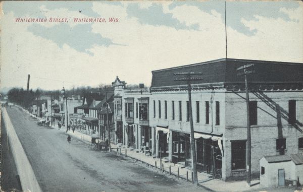 Text on front reads: "Whitewater Street, Whitewater, Wis." An elevated view of the right side of an unpaved street with sidewalks and hitching posts. Several buildings have signs. There are horse-drawn vehicles in the street.