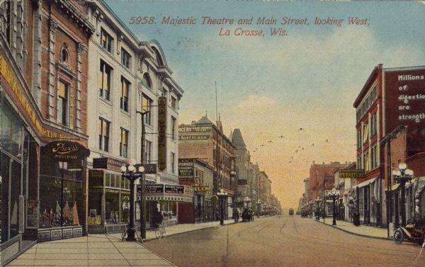 Text on front reads: "Majestic Theatre and Main Street, Looking West, La Crosse, Wis." An urban street with many buildings and businesses. Pedestrians, bicycles, automobiles, horse drawn wagons and a streetcar can be seen. Some signs read: "Pryor Studio," "Art's Drug Store,"  "Parker, Diamonds, Watches, Silverware" "Majestic," "Evening Performance," "Matinee Daily," "Furniture." 