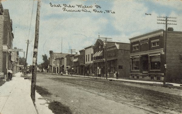 Text on front reads: "East Side Water St., Prairie Du Sac, Wis." An unpaved street with sidewalks and businesses on both sides. Some pedestrians are on the sidewalk.