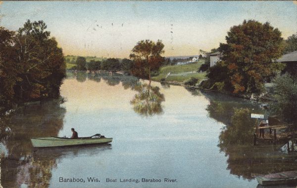 Text on front reads: "Baraboo, Wis. Boat Landing, Baraboo River." A man in a boat on the river, trees, cottages and piers are on the shorelines. Text on reverse reads: "Baraboo, Wis. was named for Jean Baribault, an early French trapper and settler. The town is beautifully built on a series of hills overlooking the Baraboo River. It is an important railway point, being the division headquarter of the North Western Line. Its manufacturing interests include woolen mills, flour mills, etc. in the center of an extensive agricultural region.