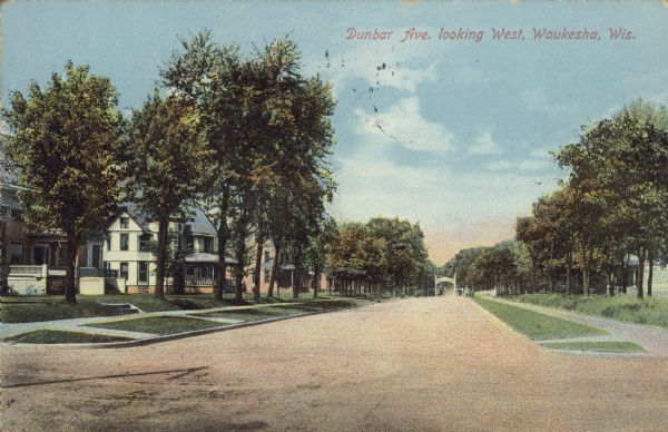 Text on front reads: "Dunbar Avenue, looking West, Waukesha, Wis." An unpaved road with curb and sidewalks in a residential neighborhood with trees. There are dwellings on both sides and a white arch and gate are visible in the far distance.