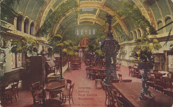 Text on front reads: "Interior, Schlitz Palm Garden, Milwaukee." A very ornately decorated space with a barrel vaulted ceiling. Skylights and arched windows provide light for palm trees and foliage. Many tables and chairs are arranged in seating groups.