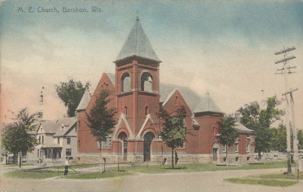 Text on front reads: "M.E. Church, Baraboo, Wis." A brick church with a corner entrance and belfry. A dwelling can be seen to the left and trees surround the building. In front is an unpaved street, sidewalks and lawns. A man is mowing the grass.