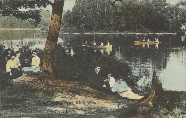 Text on front reads: "Cedar Lake near Kiel, Wis." Many people are enjoying the lake, with two groups in boats and two groups on shore. The shoreline is lined with trees.