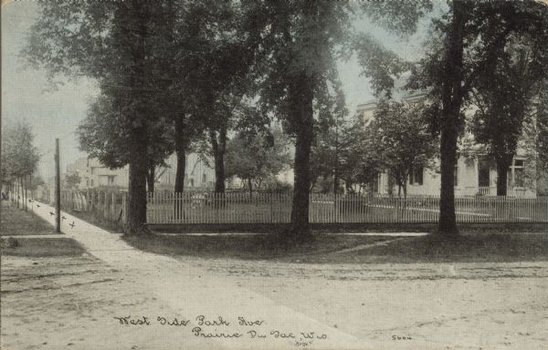 Text on front reads: "West Side Park Ave., Prairie Du Sac, Wis." Dwellings inside fenced yards on an unpaved street with sidewalks. There are many large trees in the yards and young trees along the street.