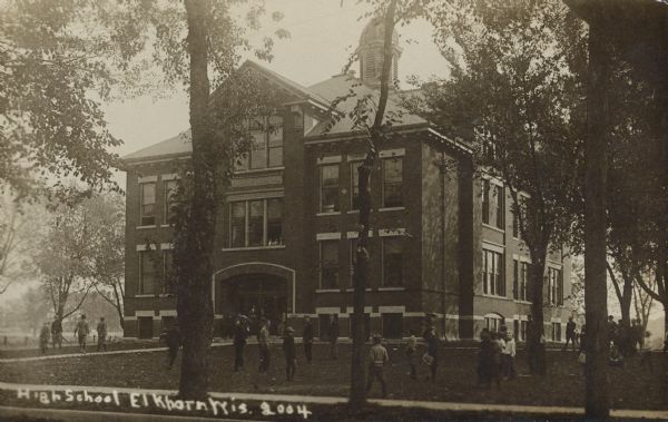 Text on front reads: "High School, Elkhorn, Wis." A three-story school with an arched entrance and belfry. Students are standing on the grounds and many mature trees surround the school.