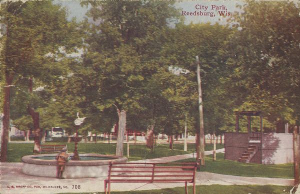 Text on front reads: "City Park, Reedsburg, Wis." A child is standing at a fountain in a park with sidewalks, trees and a bandstand.