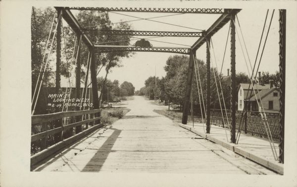 Text on front reads: "Main Street, Looking West, Rome, Wis." Unpaved street in a small town, framed by a truss bridge over the Bark River. Trees are on both sides of the street and a dwelling can be seen on the right.