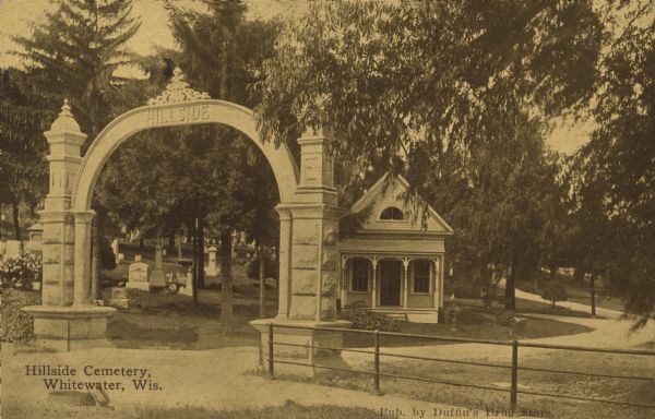Text on front reads: "Hillside Cemetery, Whitewater, Wis." View of the arched entrance to a cemetery, enclosed by a fence and filled with trees. A small house-like structure and gardens can be seen.