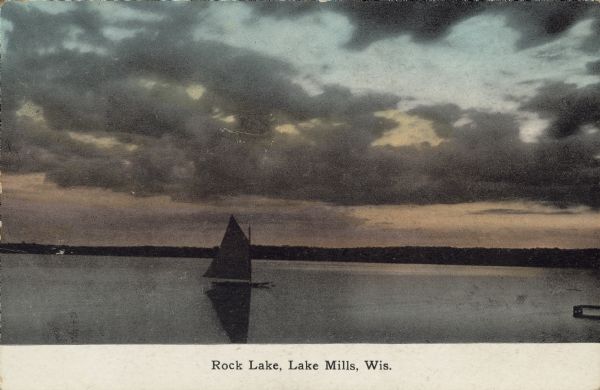 Text on front reads: "Rock Lake, Lake Mills, Wis." A sailboat on the lake at sunset. Rock Lake is a 1365 acre lake with a maximum depth of 60 feet on the West side of Lake Mills.