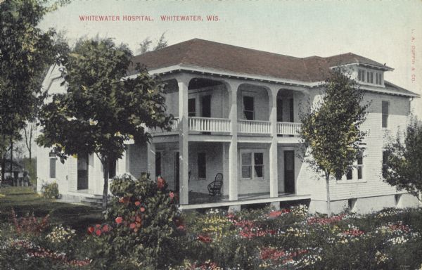 Text on front reads: "Whitewater Hospital, Whitewater, Wis." A two-story clapboard building with a porch and covered balcony above. A large flower bed is in the foreground, with trees surrounding the building. A chair is on the porch.