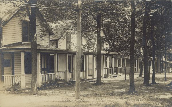 Text on front reads: "Cottage St. Evangelical Camp Grounds, Lomira, Wis." A row of cottages with porches at a church camp. The area in front of the cottages is filled with mature trees. 