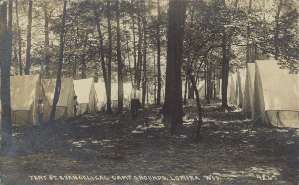 Text on front reads: "Tent St. Evangelical Camp Grounds, Lomira, Wis." A man is standing in a wooded area with rows of tents on the right and left. Two campers are standing in front of their tents.