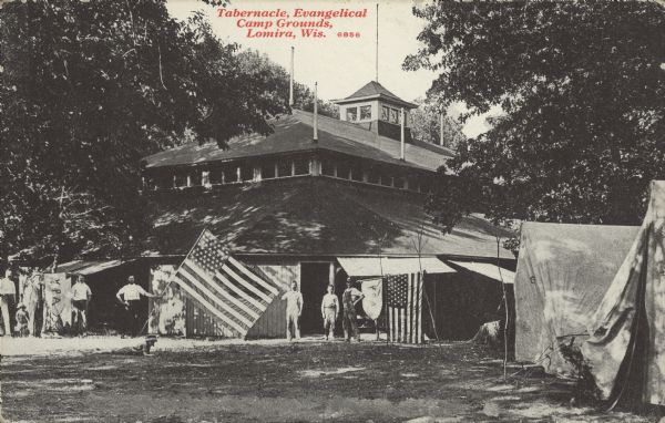 Text on front reads: "Tabernacle, Evangelical Camp Grounds, Lomira, Wis." Adults and children pose in front of the Tabernacle at the Evangelical Camp Grounds with American flags. There are tents on the right and mature trees all around.