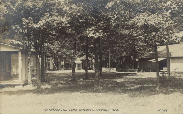 Text on front reads: "Evangelical Camp Grounds, Lomira, Wis." A row of cottages with porches at a church camp. A large building is on the right. The area between is filled with mature trees and a bench.