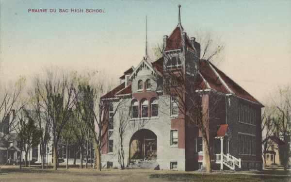 Text on front reads: "Prairie Du Sac High School". A multistory school building with an arched entrance and arched windows. A church is on the left. It became the Sauk Prairie Junior High and was destroyed in a fire in 1965.