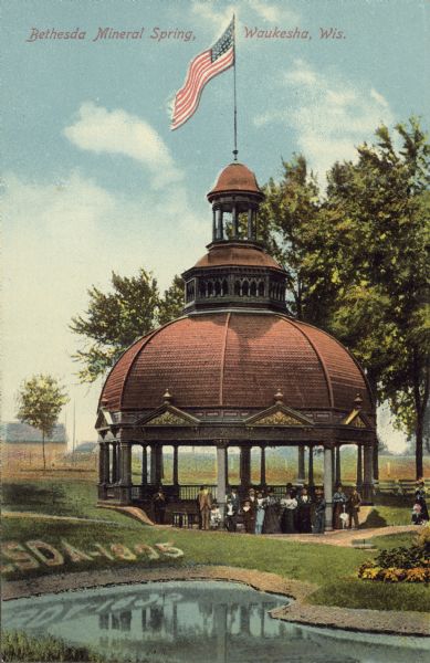 Text on front reads: "Bethesda Mineral Spring, Waukesha, Wis." A group of people in the pavilion at Bethesda Mineral Spring. An American flag is flying at the top of the domed pavilion. Between 1868 and 1918, 60 mineral springs were located here. People would travel here to "enjoy the waters."