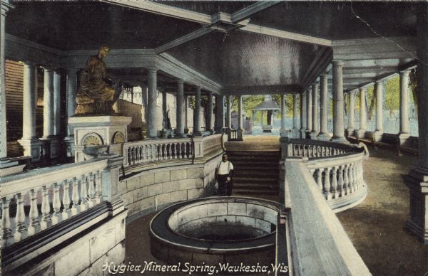 Text on front reads: "Hygiea Mineral Spring, Waukesha, Wis." The spring is shown inside a pavilion and surrounded by stone walls and balustrades. There is a statue on a column on the left. A man is posing behind the spring. Between 1868 and 1918, 60 mineral springs were located here. People would travel here to "enjoy the waters."