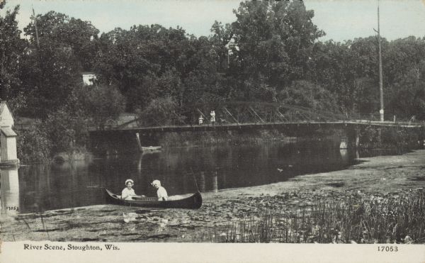 Text on front reads: "River Scene, Stoughton, Wis." Two women in a canoe on the Yahara River near wetlands foliage, with two people on the pedestrian bridge in the background. A building is on the left on the opposite shoreline which is covered with trees.