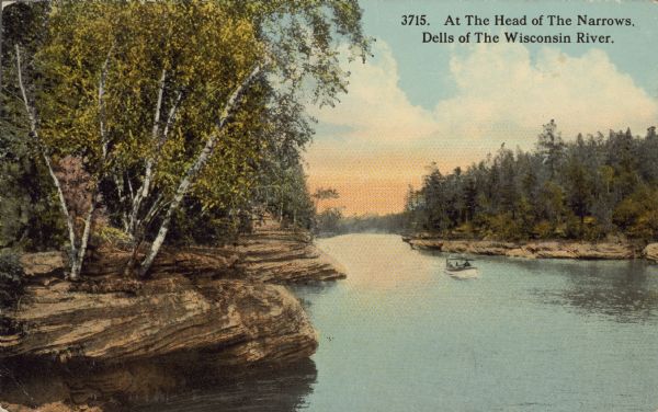Text on front reads: "At The Head of The Narrows, Dells of The Wisconsin River." A channel of the Wisconsin River that is 52 feet wide and 100 feet deep. The shoreline is rock formations and trees, and an excursion boat is on the water.