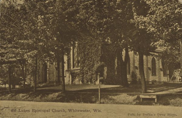 Text on front reads: "St. Luke's Episcopal Church, Whitewater, Wis." Ground level view of a church, mostly obscured by mature trees.