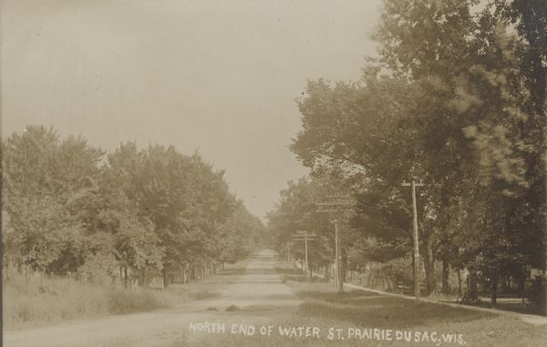 Text on front reads: "North End of Water Street, Prairie du Sac, Wis." View of a long unpaved street, with a sidewalk, utility poles and trees. Fences are along both sides of the street, with a bicycle parked against a tree near the sidewalk on the right.