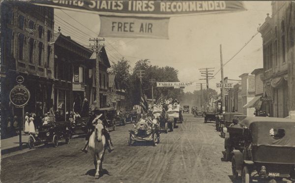 Handwriting on reverse reads: "Uncle John Doll leading 4th of July Parade in 1915." A man on a horse leads a parade of automobiles, trucks and tractors down an unpaved main street. Banners are strung overhead and many buildings have signs. Banners read: "United States Tires Recommended, Free Air" and "Ford Garage." The giant watch on the left reads: F.S. Eberhart, City Jeweler." Vehicles are parked on both sides of the street.