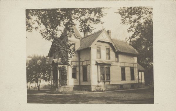 Handwriting on reverse reads: "House across the street at 485 Broadway, Pra. du Sac." A two-story clapboard home with an attic and basement. A tower is above the entrance porch and the house has decorative elements. A sidewalk runs around the house which is surrounded by trees.
