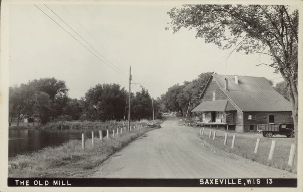 Text on front reads: "The Old Mill, Saxeville, Wis." An tree-lined, unpaved road with white guard posts vanishes into the distance. The mill is on the right and the millpond, on the Pine River, is on the left. A truck is in the parking lot.