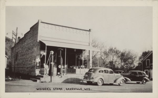 Text on front reads: "Weiser's Store, Saxeville, Wis." A brick building with a decorative storefront has a raised concrete platform and gasoline pumps in front. One automobile is parked in front, two more on the side. Another building is on the right.