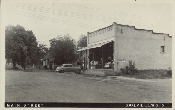 Text on front reads: "Main Street, Saxeville, Wis." Weiser's Store, a brick building with a decorative storefront has a raised concrete platform and gasoline pumps in front. An automobile is parked at the corner. A house and trees are in the background on the left.