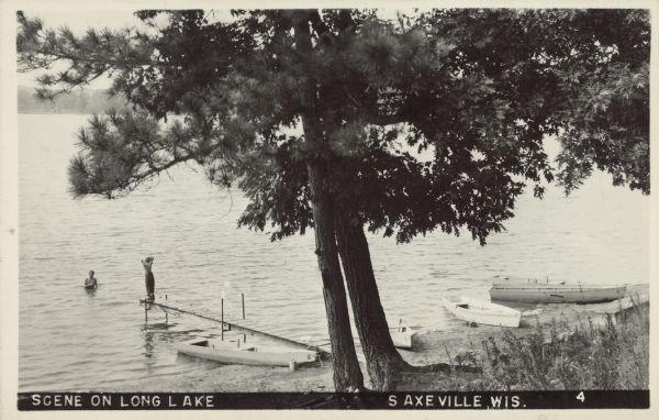 Text on front reads: "Scene on Long Lake, Saxeville, Wis." A dock is on the shore of a lake with a large tree overhead. One person is on the dock, another is in the water. Four rowboats are pulled up on the shore.
