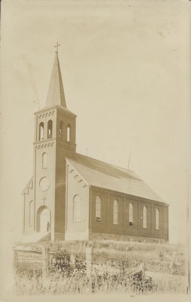 Handwritten on reverse: "Saint Ann's Catholic Church, Saxon, Wis." Built in 1909, the church is still serving the needs of the community today. In recent years, several projects have taken place to restore the building. A man is standing in front of the entrance.