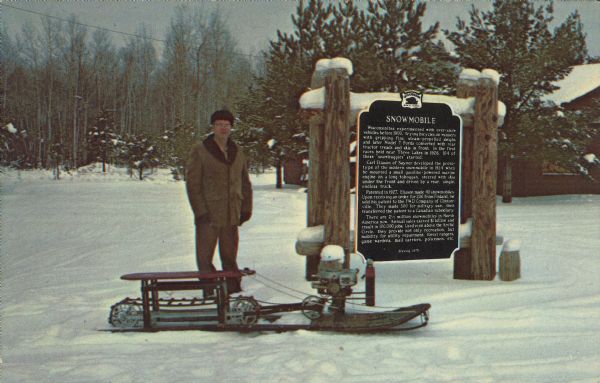 Text on reverse reads: "World's First Snowmobile, Sayner, Wisconsin. Invented in 1924 by Carl Eliason of Sayner (pictured). This machine is liquid cooled, has slide rails, is powered by 2 1/2 horsepower, and can reach 20 mph. This machine is displayed at Carl Eliason & Co. or the museum in Sayner." He is standing next to the Wisconsin Historical Marker #212, erected in 1975, honoring him for his invention. There is a building and trees in the background, covered with snow.