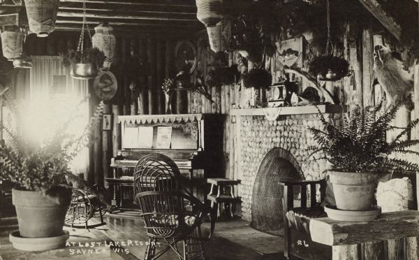 Text on front reads: "At Lost Lake Resort, Sayner, Wis." The lodge at a Northern Wisconsin resort is built of logs. Chairs are arranged around the fireplace, with a piano in the corner. It is decorated with many items, including plants, taxidermy, lanterns and pictures.